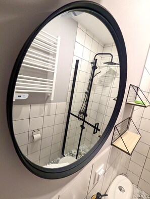 Une salle de bain avec un miroir rond, une douche équipée d’un pommeau de douche à main, des toilettes et un porte-serviettes.