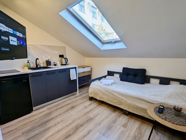 Une chambre à coucher de taille réduite, dotée d’une lucarne, d’un lit recouvert d’une couette blanche, d’une petite table de chevet et offrant une vue sur l’extérieur grâce à la lucarne.
