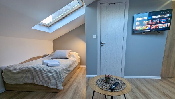 A bedroom with a bed, a small round table, a TV mounted on the wall, a door, and a skylight.