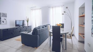 Ein modernes Wohnzimmer mit einer Couch, einem Esstisch und Stühlen. Ein Fernseher steht auf einer Kommode, und auf dem Tisch steht eine Pflanze.