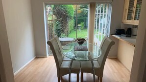 Un coin repas avec une table en verre et quatre chaises, un petit saladier rempli de fruits, et une vue sur un jardin par une porte-fenêtre coulissante.