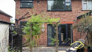 Une maison à deux étages, construite en briques, avec une porte d’entrée, des fenêtres et un petit jardin agrémenté de plantes et d’une chaise.