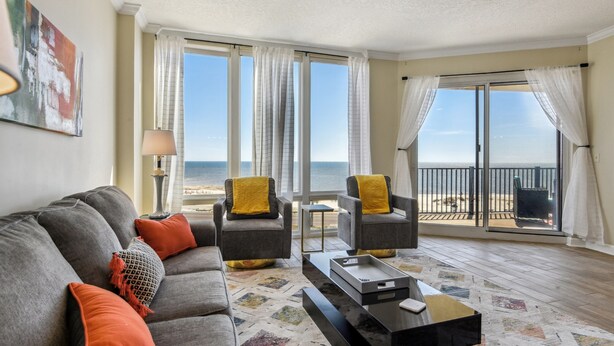 Sala de estar com sofá cinza, duas poltronas, mesa de centro e vista para a praia através de grandes janelas.