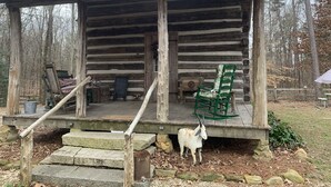Een rustieke houten chalet met een veranda, een groene stoel, een geit en een bord dat aangeeft dat er kip op het menu staat.