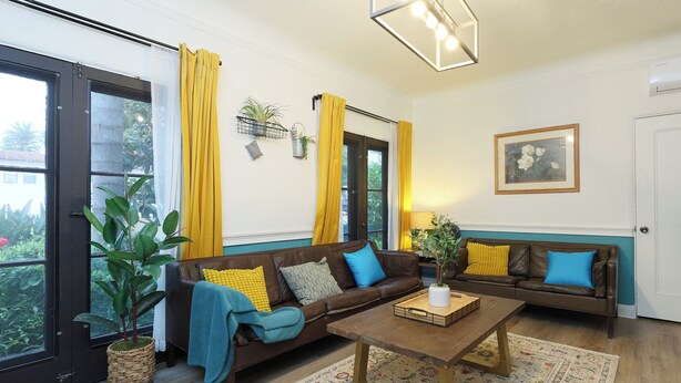 Ein Wohnzimmer mit einer braunen Ledersofa, einem Holztisch und einem gemusterten Teppich. Gelbe Vorhänge, ein Blumentopf und ein gerahmtes Bild sind an der Wand zu sehen.