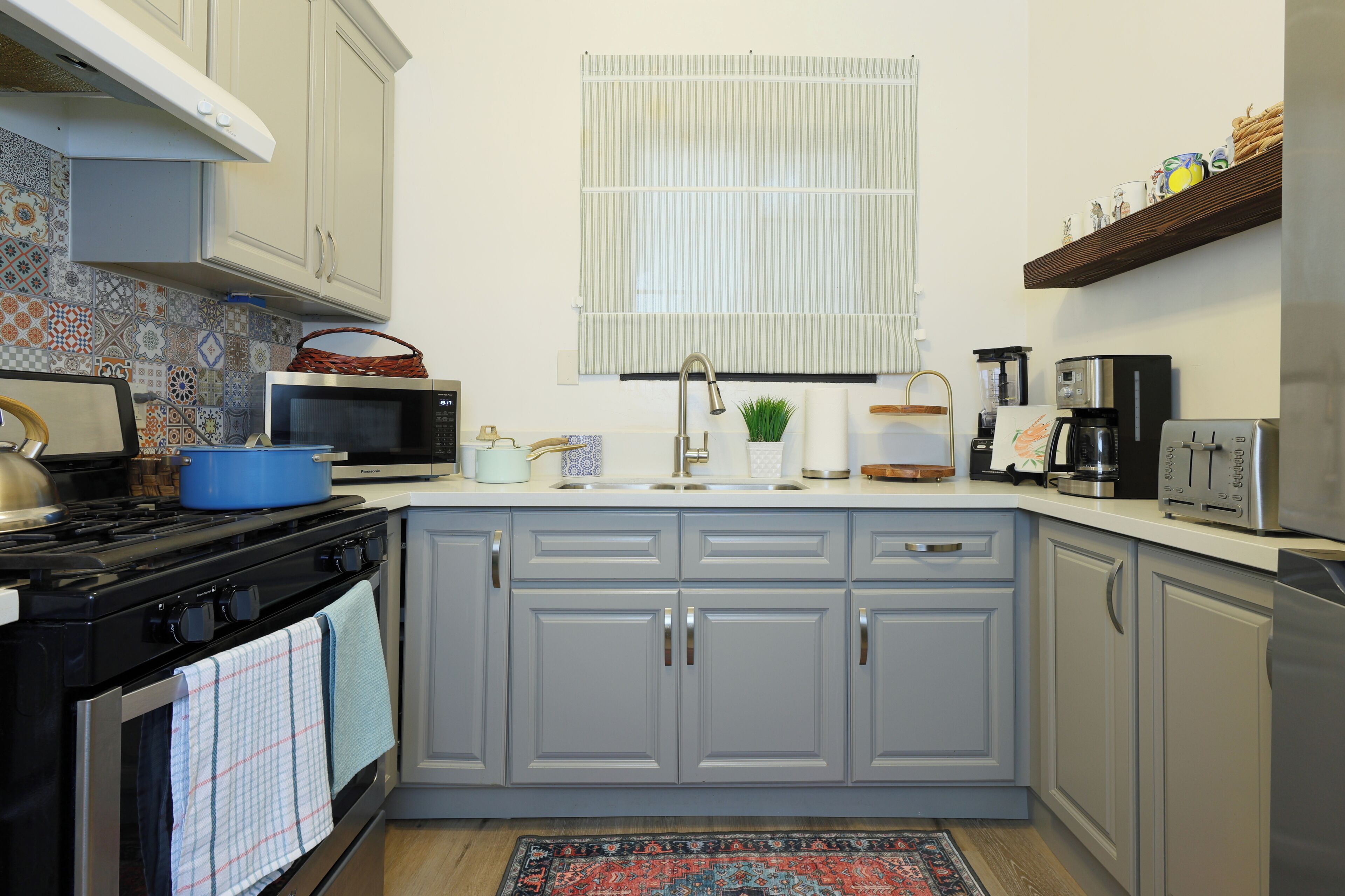 A kitchen with grey cabinets, a stove, a microwave, a sink, and a window with blinds.