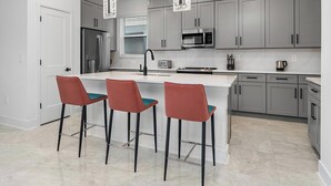 A modern kitchen with grey cabinets, a central island with bar stools, stainless steel appliances, and pendant lighting. - The Mystical | Ocean-lover's home w PVT pool, submarine theme BR, near waterpark (Orlando)