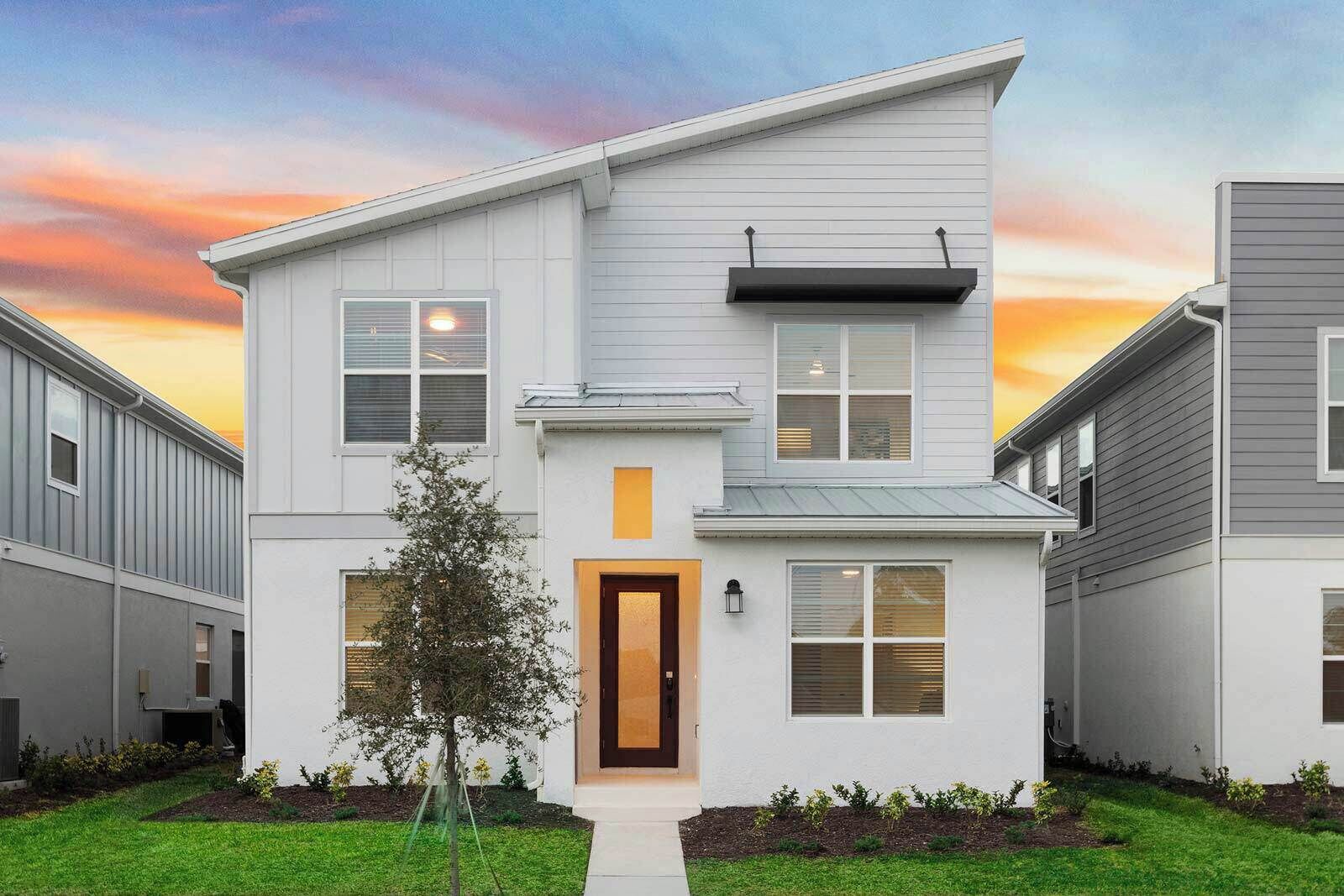 A modern two-story house with a central entrance, a small tree in the front yard, and a sunset sky.