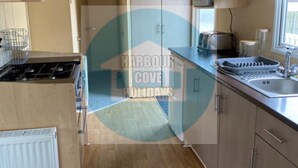 A compact kitchen with a gas stove, microwave, and sink. A 'No Smoking' sign is visible. The floor is wooden, and the kitchen has light-colored cabinets. - Affordable Caravan Holidays, Butlins Minehead (Minehead)