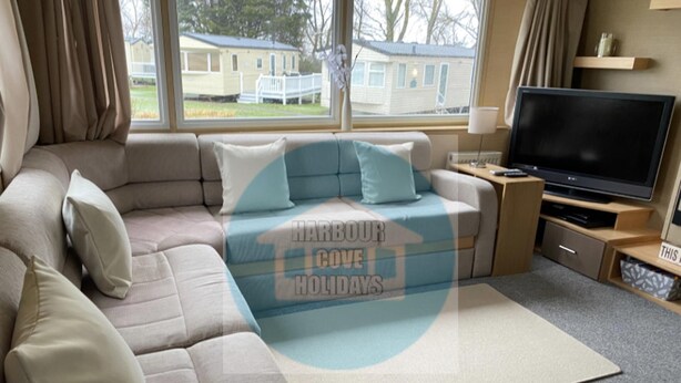 A cozy living room with a sectional sofa, a flat-screen TV, and a view of a residential area through large windows. - Affordable Caravan Holidays, Butlins Minehead (Minehead)