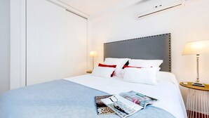 A neatly made bed with a light blue bedspread, white pillows, and a grey headboard. There are two bedside tables with lamps, and a magazine is placed on the bed. - Genteel Home Placentines (Seville)