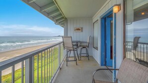A balcony with a view of the beach, a table with chairs, and a blue door. - Oceanfront Condo Amazing Views, Charming Nautical Theme, Covered Deck (Newport)