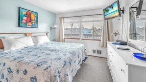A hotel room with a bed, a desk, a TV, and a window with a view of houses. - Oceanfront Condo Amazing Views, Charming Nautical Theme, Covered Deck (Newport)