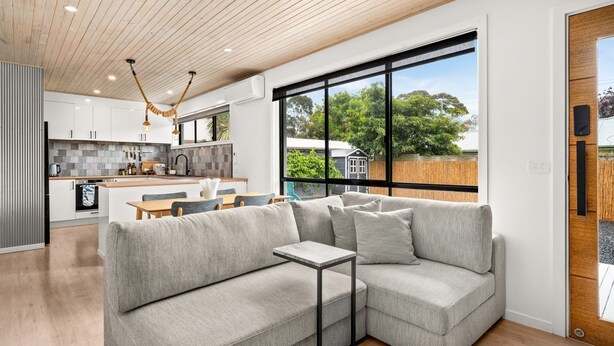 Een moderne woonkamer met een hoekbank, een eethoek en een keuken in de achtergrond, plus een groot raam met uitzicht op de omgeving.