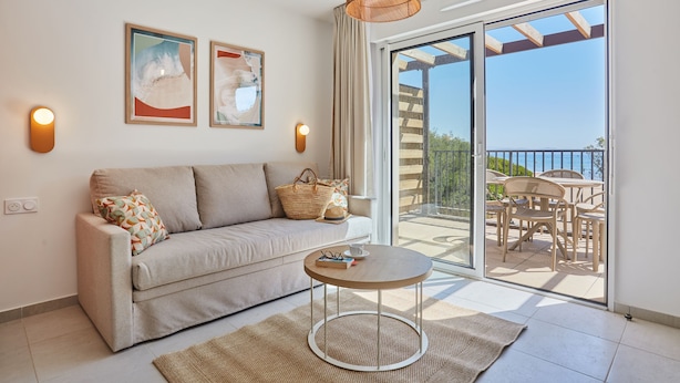 A modern living room with a sofa, a round table, and a balcony with outdoor furniture. - Résidence Odalys Riviera la Presqu'Ile (Hyères)