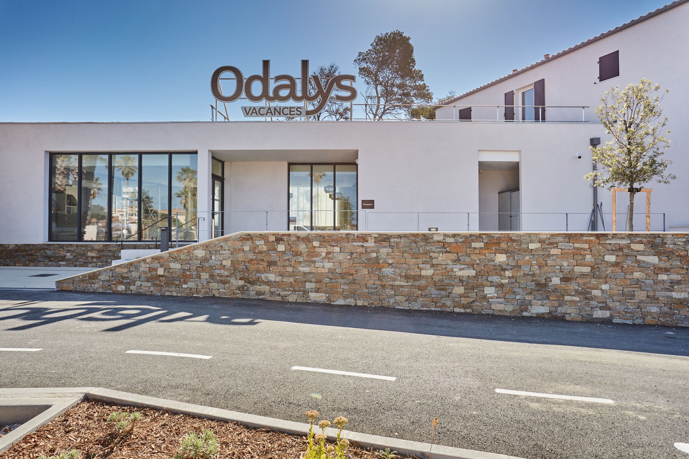 Un bâtiment moderne avec une façade en pierre, de grandes fenêtres et une enseigne indiquant « Odaly's Vacances ».