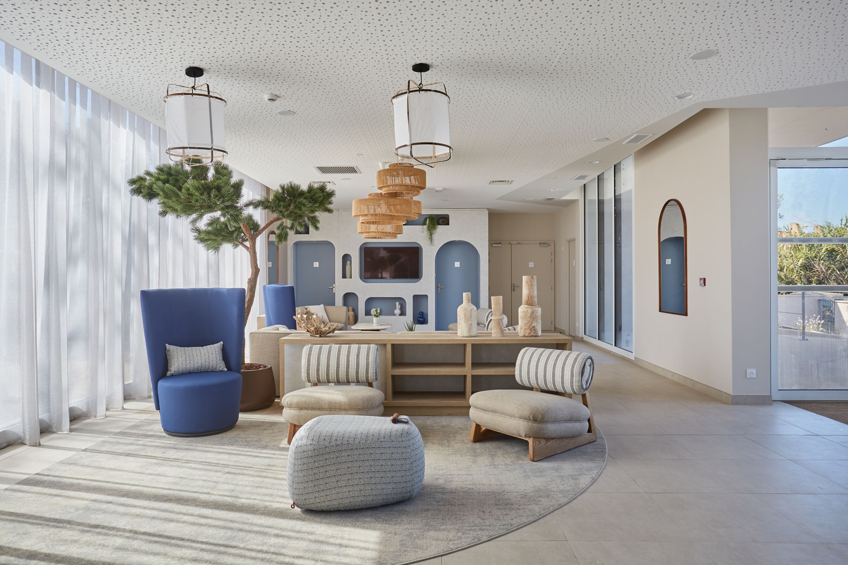 Uma sala de estar moderna com uma grande poltrona azul, um banquinho redondo e uma mesa de centro de madeira. Há duas luminárias pendentes, um espelho grande e um vaso de plantas.