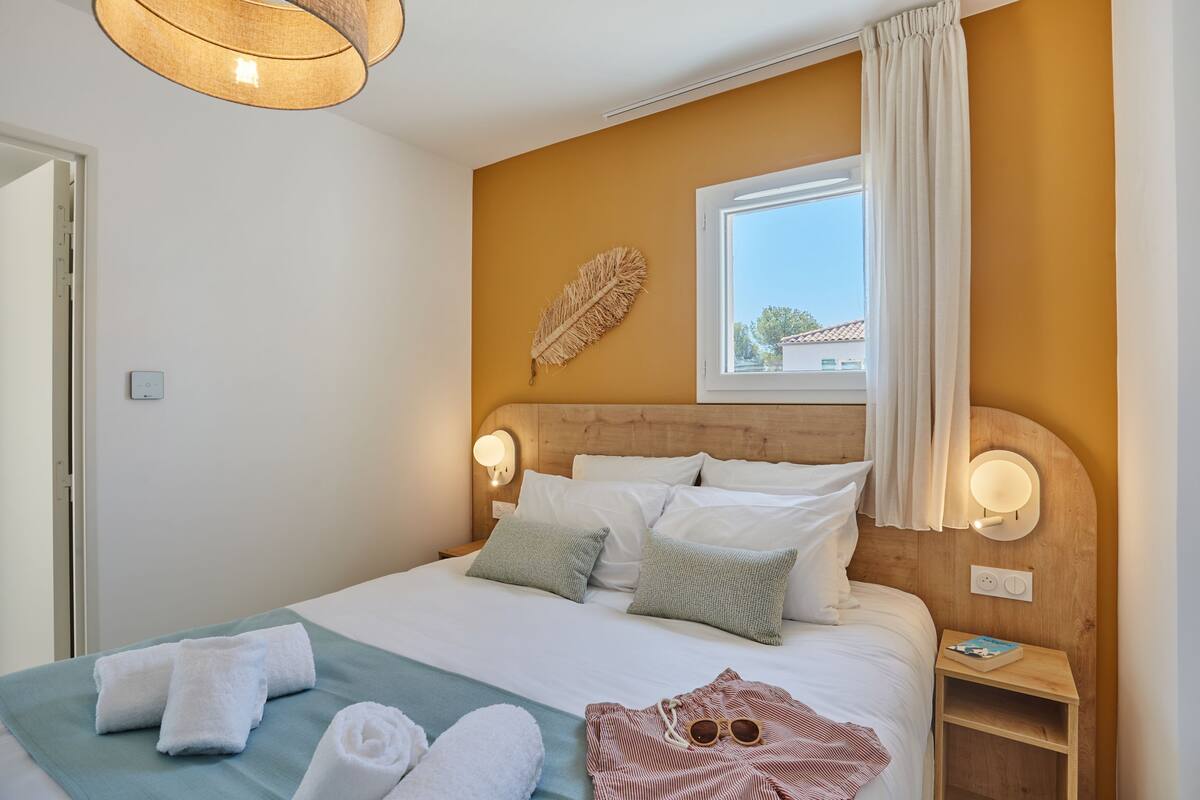 A neatly made bed with white and green pillows, a bedside table with a book, and a wall-mounted feather decoration.