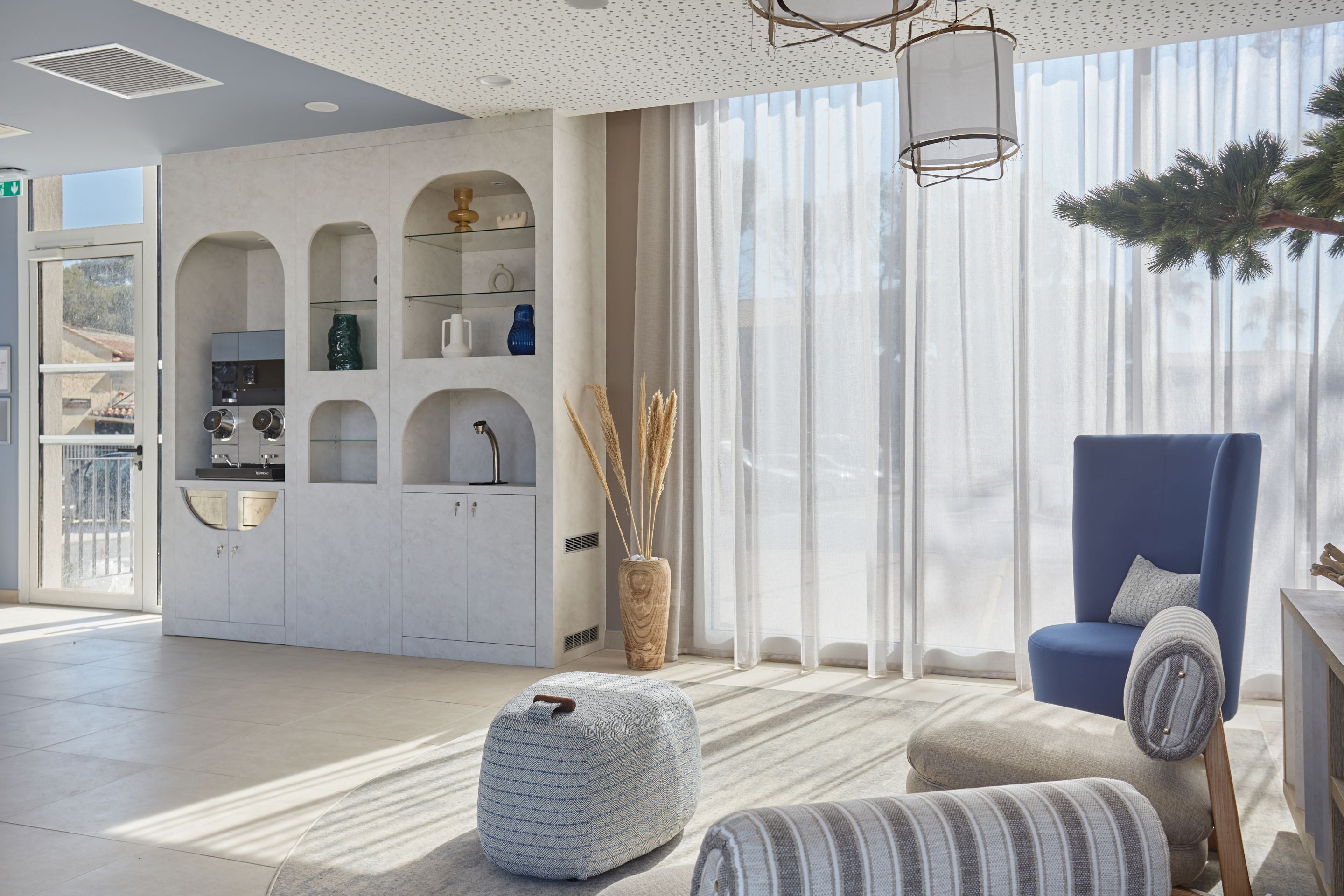 A modern living room with a blue armchair, a patterned ottoman, and a kitchen area with white cabinetry and a window with sheer curtains.