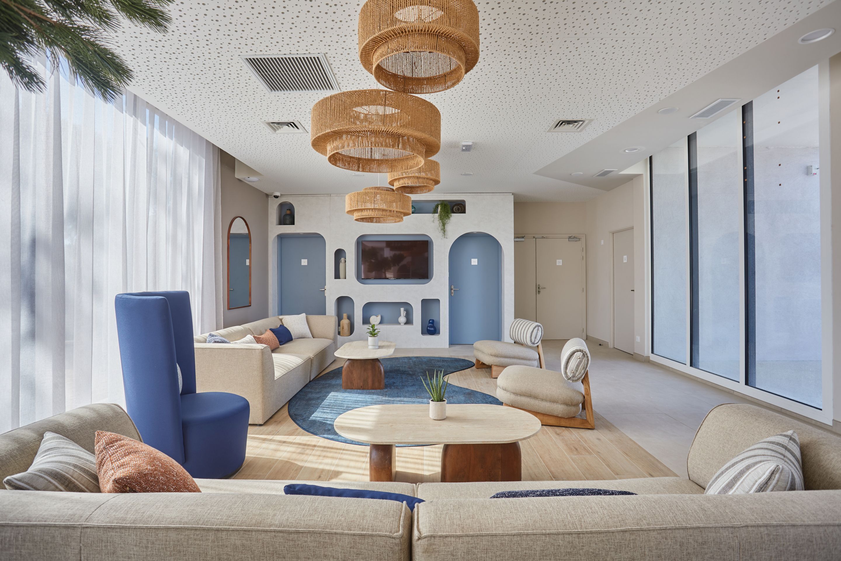 Een moderne woonkamer met een grote bank, een salontafel en een televisie. Er hangen rieten lampen aan het plafond en bevindt zich een groot raam aan de rechterkant.