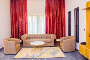 Una sala de estar con un sofá color crema, dos sillones y una mesita de centro. Se ven cortinas rojas y una ventana.