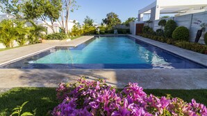 A rectangular swimming pool with clear blue water, surrounded by a concrete deck and lush greenery, including purple flowers and various trees. - Modern 4-BR Villa in Gated Community (Punta Cana)