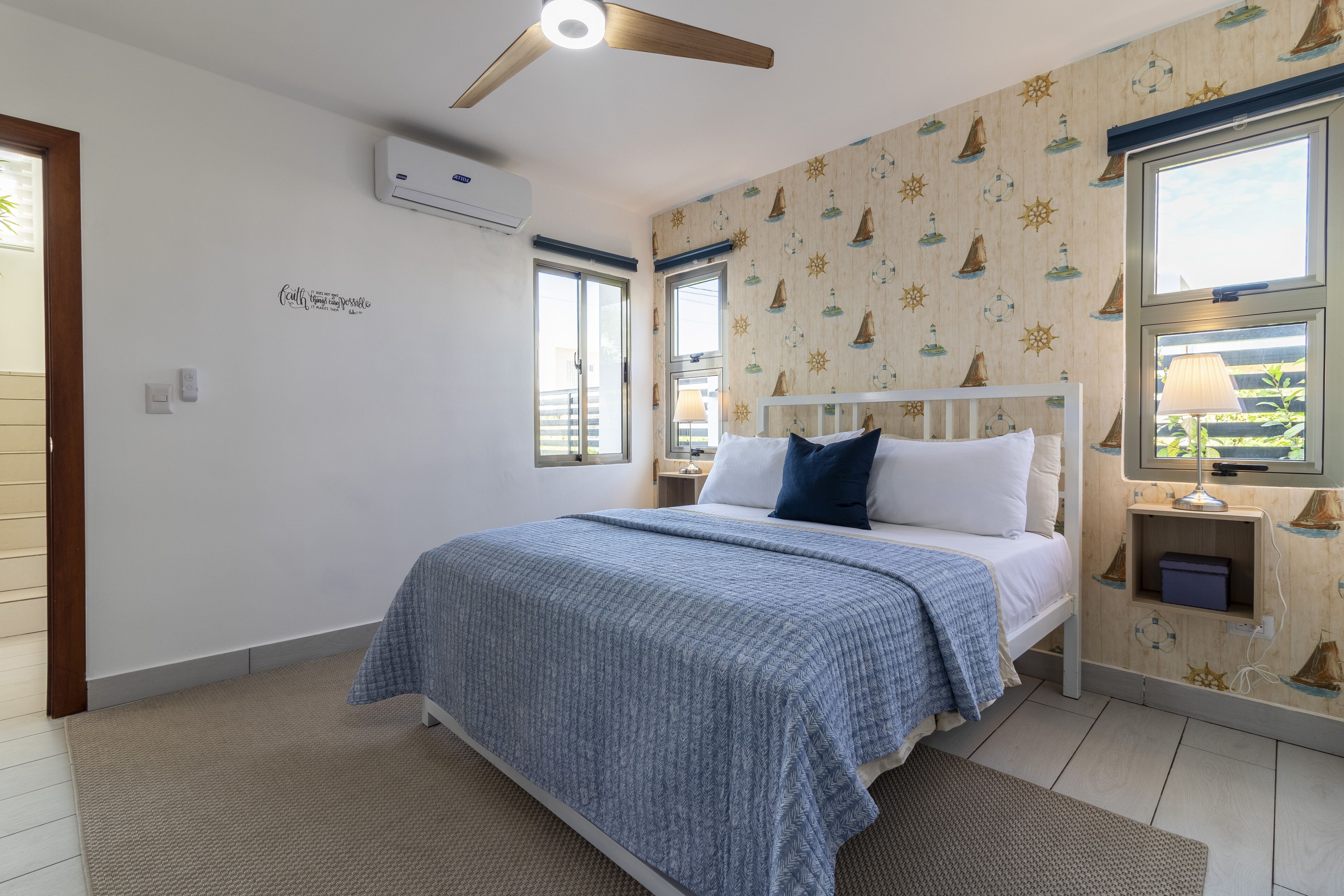A bedroom with a bed, a ceiling fan, a window, and a wall with nautical-themed wallpaper.