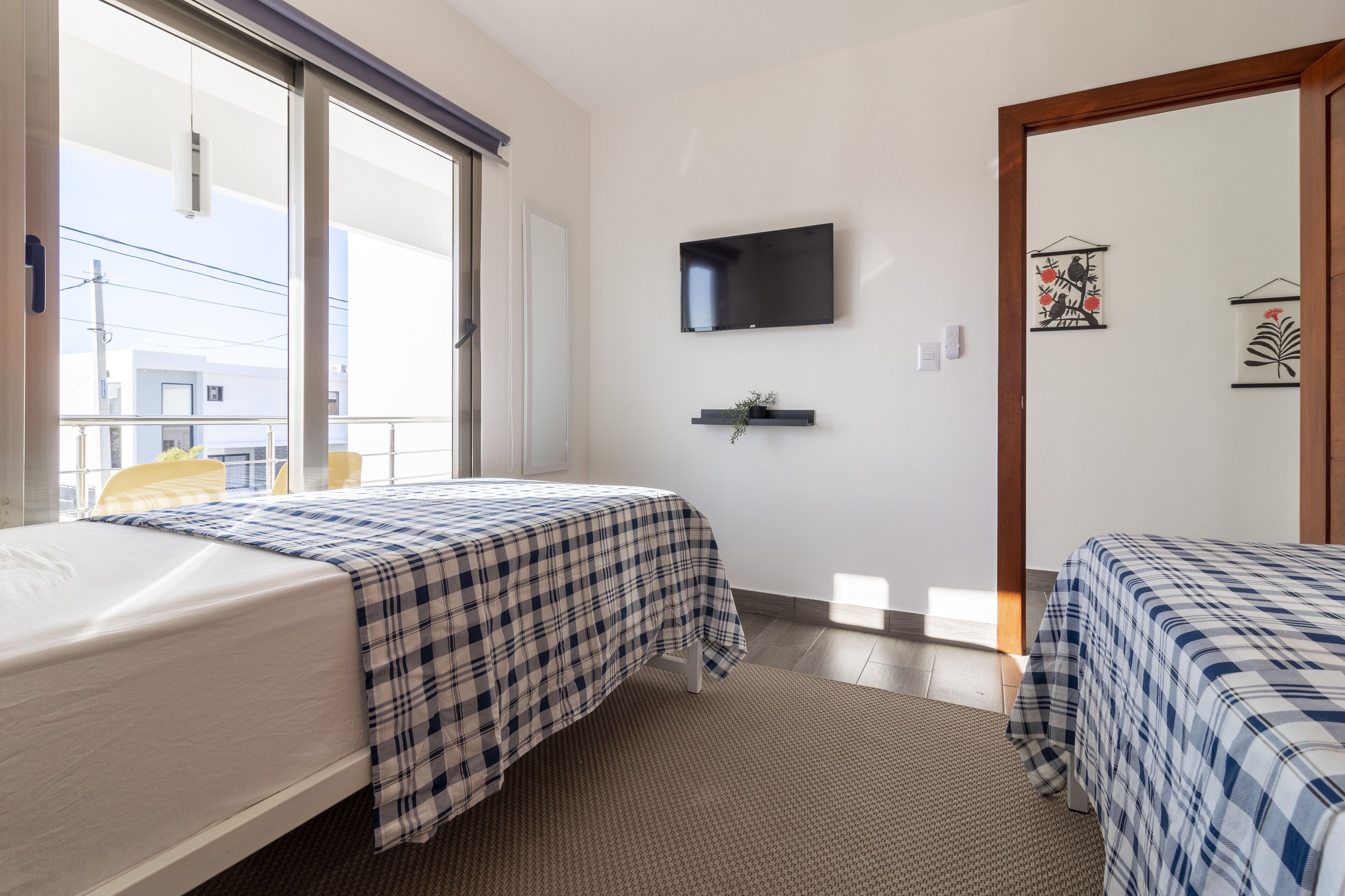 A hotel room with two beds, a TV mounted on the wall, a small shelf with a plant, and a window with a view of buildings outside.