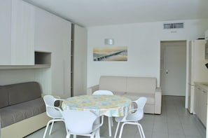 A compact living space with a sofa, a dining table with chairs, a painting on the wall, and a door leading to another room. - Charming Studio Apartment With Garden View (San Michele al Tagliamento)