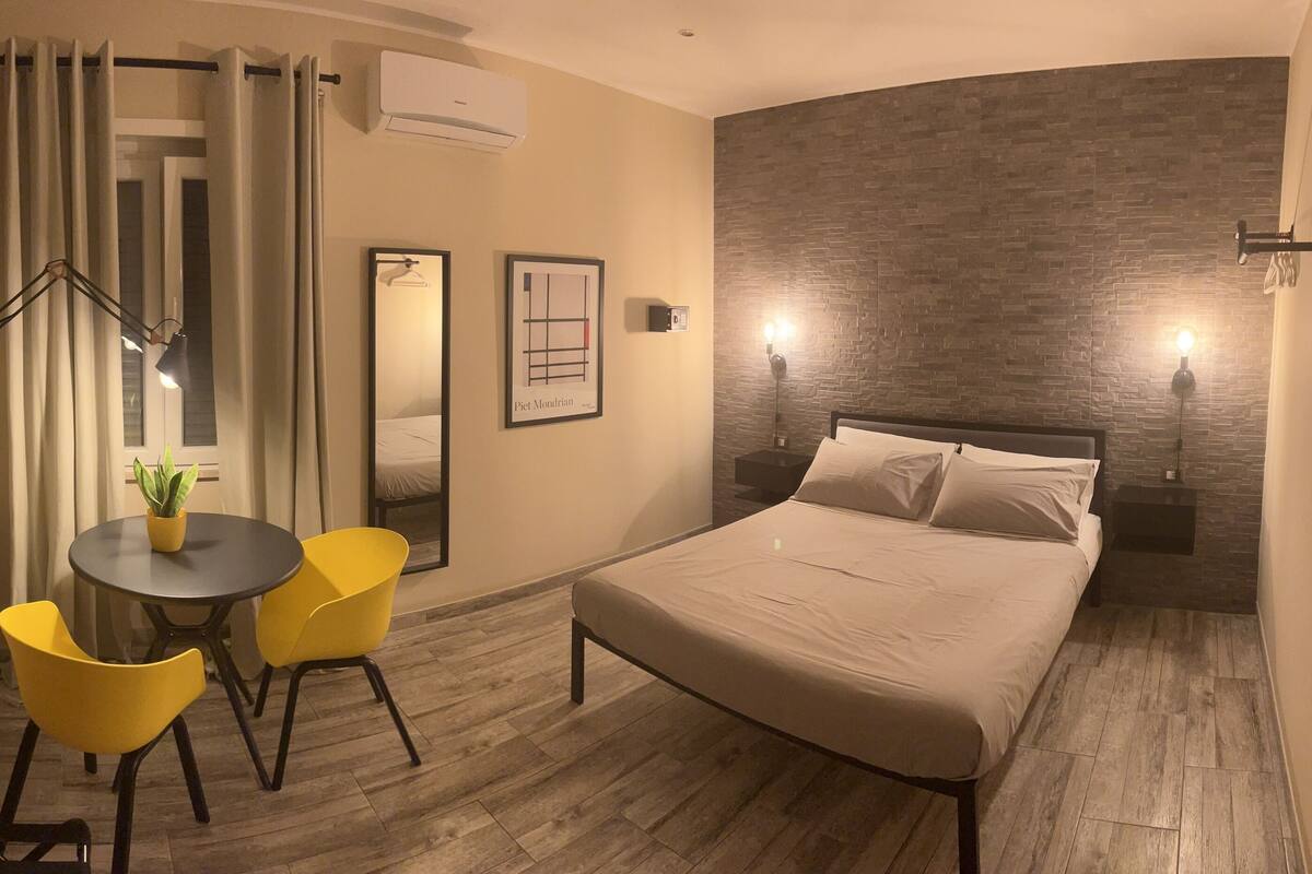 A hotel room with a bed, a desk with a chair, a potted plant, and a wall-mounted TV.