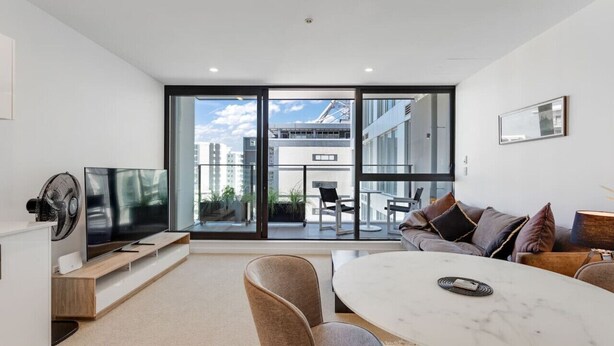 A modern living room with a large TV, comfortable seating, and a view of a cityscape through large windows. - The CityScape Apartment with Wi-Fi and Balcony (Auckland CBD)