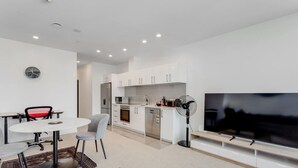 A modern kitchen with white cabinets, a built-in oven, and a microwave. There is a dining area with a round table and chairs, and a large flat-screen TV mounted on the wall. - The CityScape Apartment with Wi-Fi and Balcony (Auckland CBD)