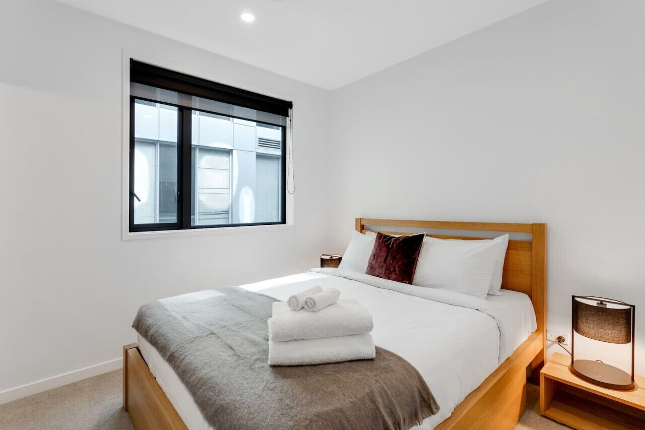 Une chambre d’hôtel moderne avec un lit en bois, du linge de lit blanc, une fenêtre avec des stores et une lampe de chevet.