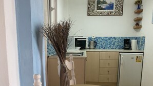 A dining area with a round wooden table, white chairs, a vase with dried plants, a microwave, a coffee maker, and a refrigerator. - Panorama Notos (Corfu)