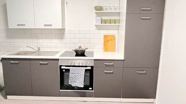 Cocina compacta con horno integrado, estufa y lavamanos. Hay armarios con manijas y un estante sobre el lavamanos.