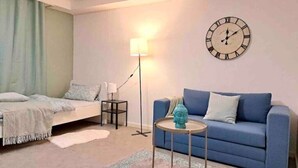 Una habitación con una cama, un sofá azul, una mesita con un adorno, una lámpara de pie y un reloj de pared.