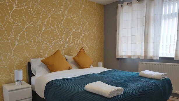 A neatly made bed with a teal bedspread and two mustard pillows, a bedside table with a lamp, and a window with sheer curtains.