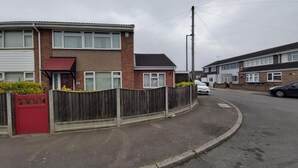 A residential street with houses, a red gate, and a white car parked on the side.