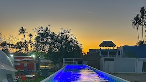Una piscina al aire libre con tobogán de agua, rodeada de palmeras y un atardecer de fondo.