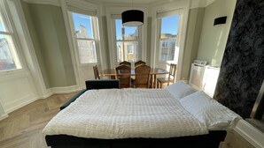 A bedroom with a large bed, a dining table, chairs, and a view of buildings through the windows. - Regency Style Spacious Seaview Apartment. Sleeps up to 6 (Brighton and Hove)