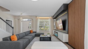 Un salon moderne avec un canapé d’angle gris, un pouf blanc, une table basse noire et un téléviseur à écran plat fixé à un mur en bois.