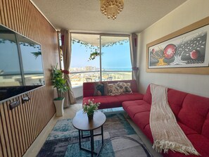 Ein Wohnzimmer mit einem roten Sofa, einem kleinen Tisch mit einer Pflanze, einem Bild an der Wand und Blick auf Stadt und Meer durch große Fenster.