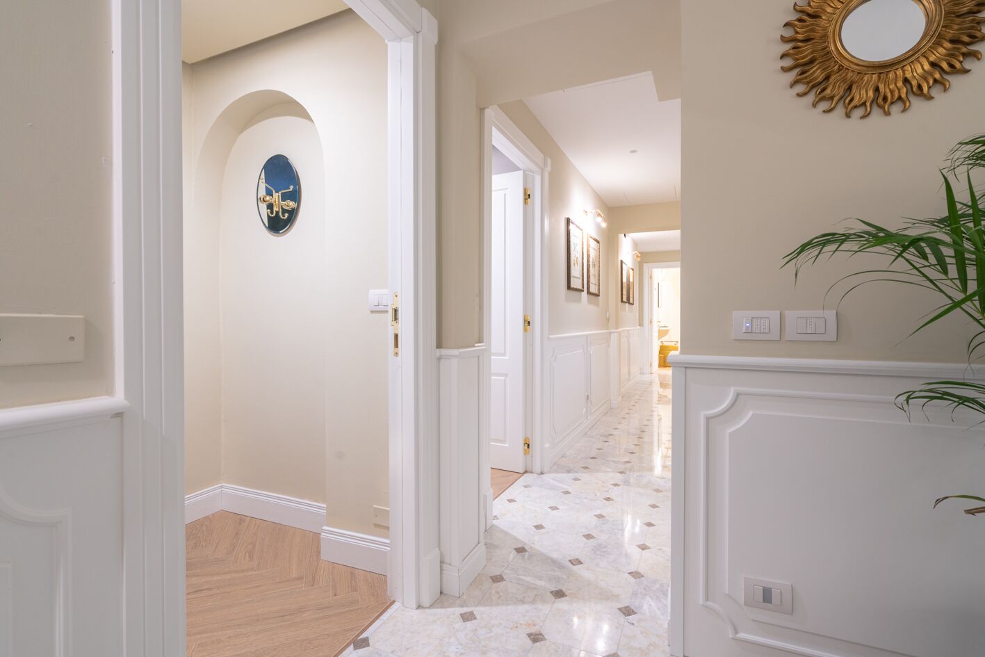A hallway with a marble floor, white walls, and a sunburst mirror.