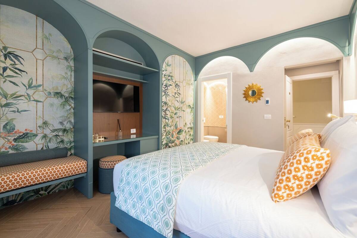 A bedroom with a bed, a bench, a TV, and a bathroom visible through an archway.