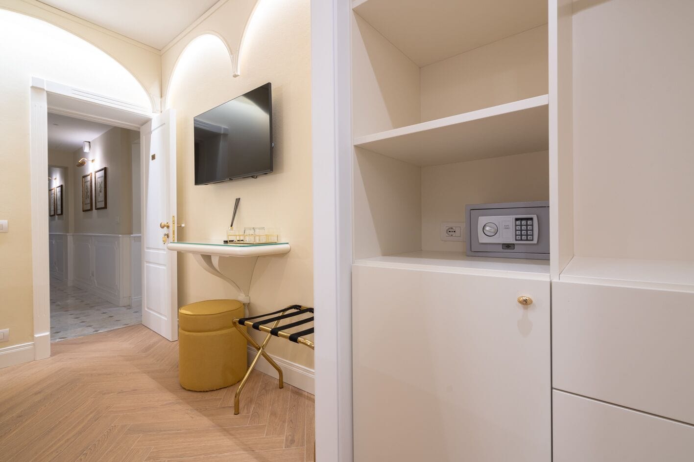 A hallway with a flat-screen TV mounted on the wall, a small vanity with a mirror, and a security safe on a shelf.