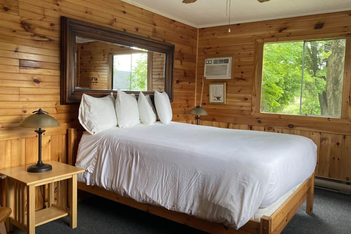 A wooden cabin room with a bed, two bedside tables, a mirror, and a window with a view of trees.