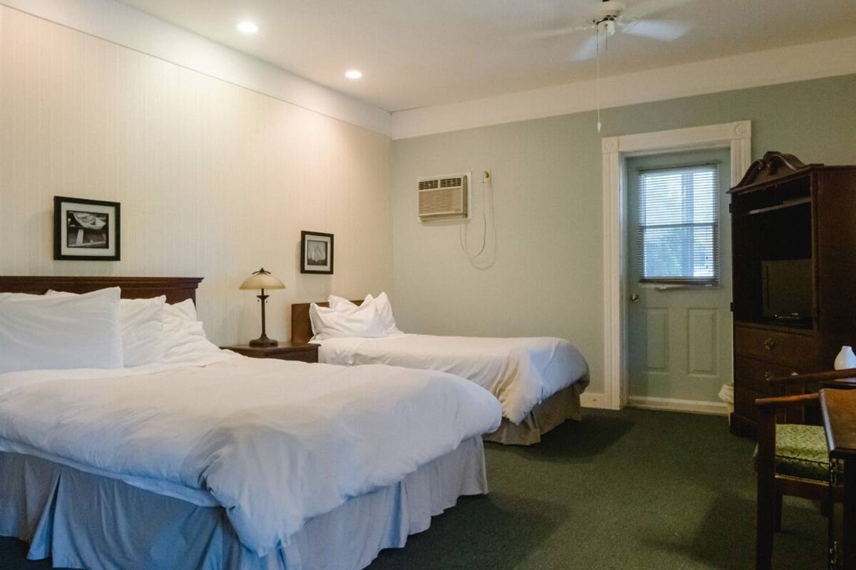 A hotel room with two beds, a desk, a chair, a ceiling fan, an air conditioning unit, and a door with a window.