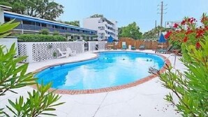 A swimming pool surrounded by a white fence, with a building in the background and greenery around.