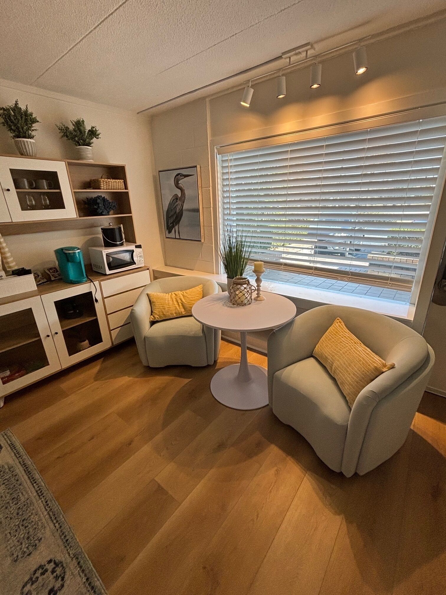 A modern living room with a round table, two armchairs, a sofa, a cabinet with glass doors, and a window with blinds.