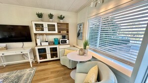 A modern living room with a flat-screen TV, a white desk, a sofa, and a dining table. There are cabinets with glass doors and a window with blinds.